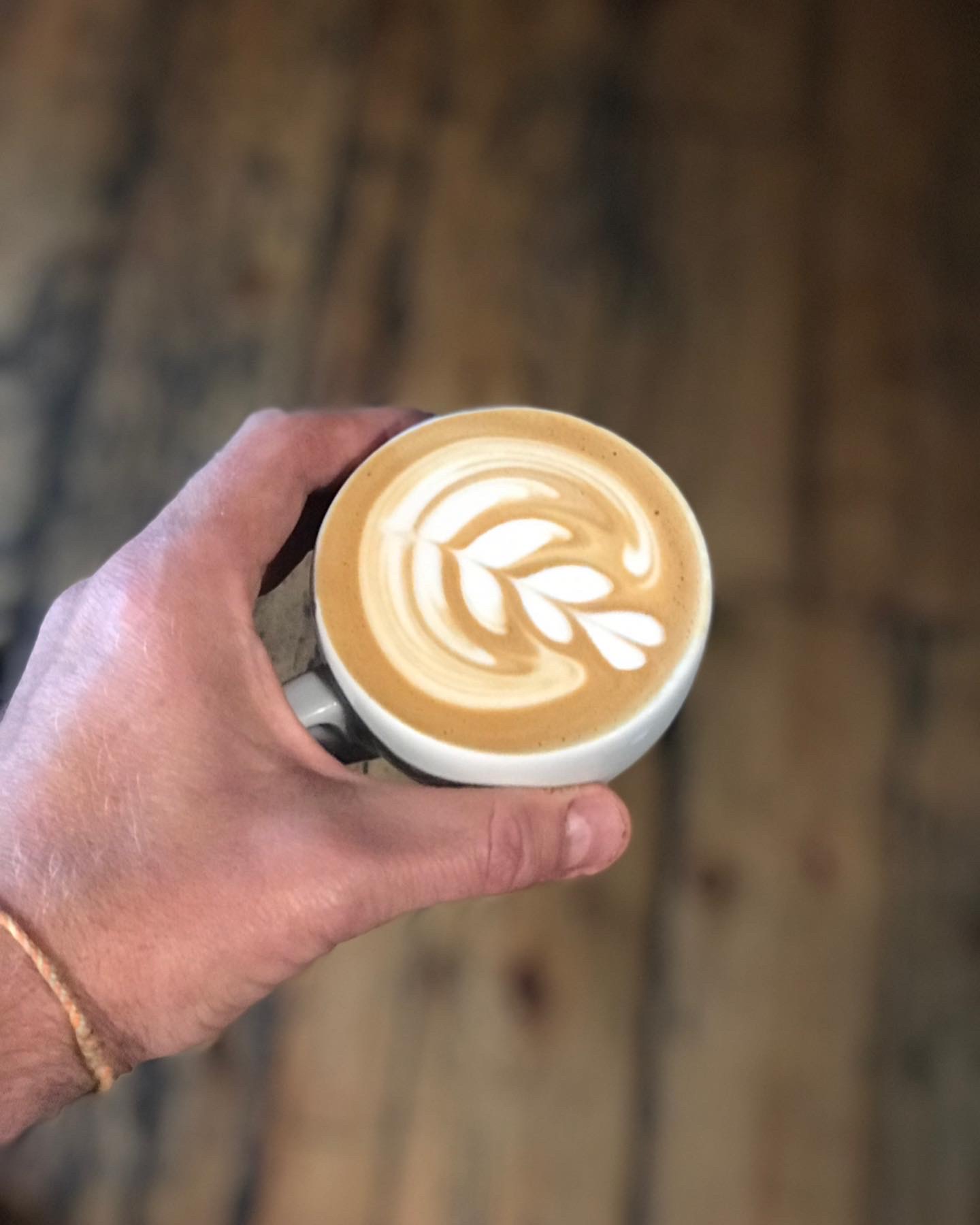 Hand holding a flat white with a crisp multi-leaf rosetta over reclaimed wooden floorboards